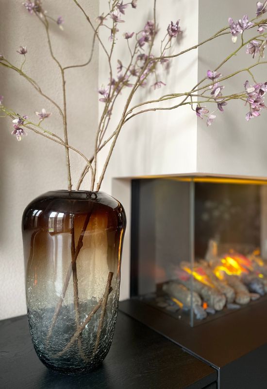 Bruin grijze vaas met paarse kunstbloemen naast open haard