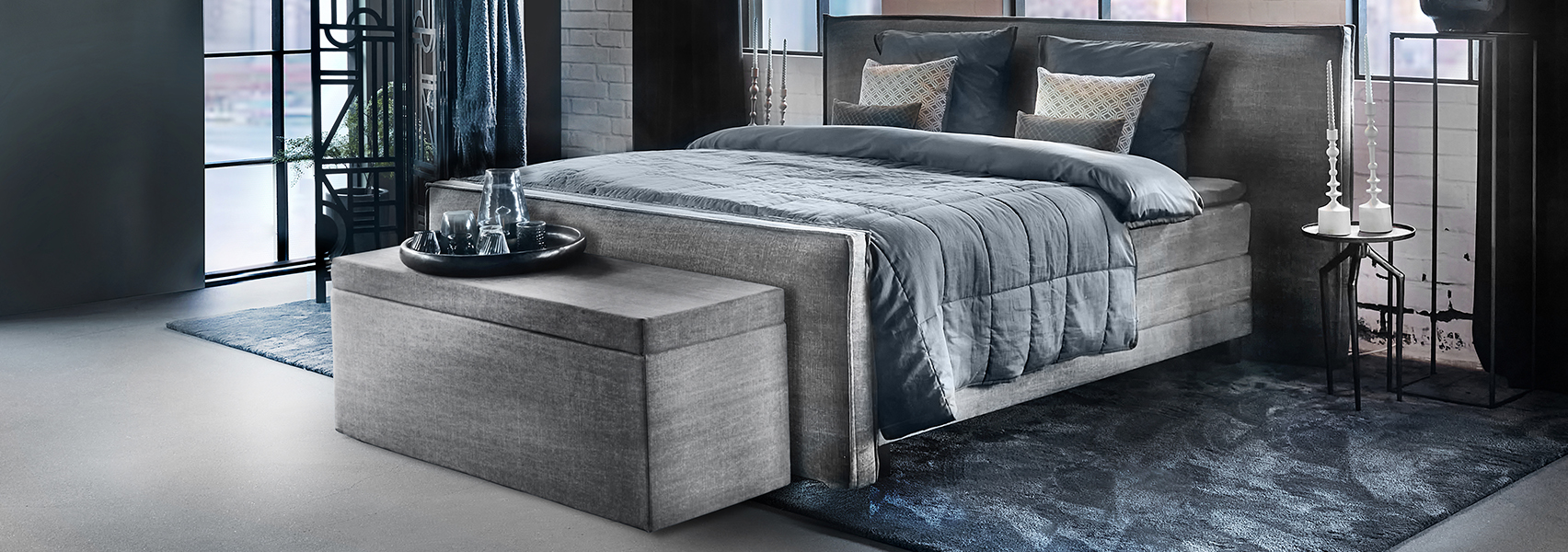 Grijze boxspring met bijpassende dekenkist blauw beddengoed en laagpolig blauw vloerkleed